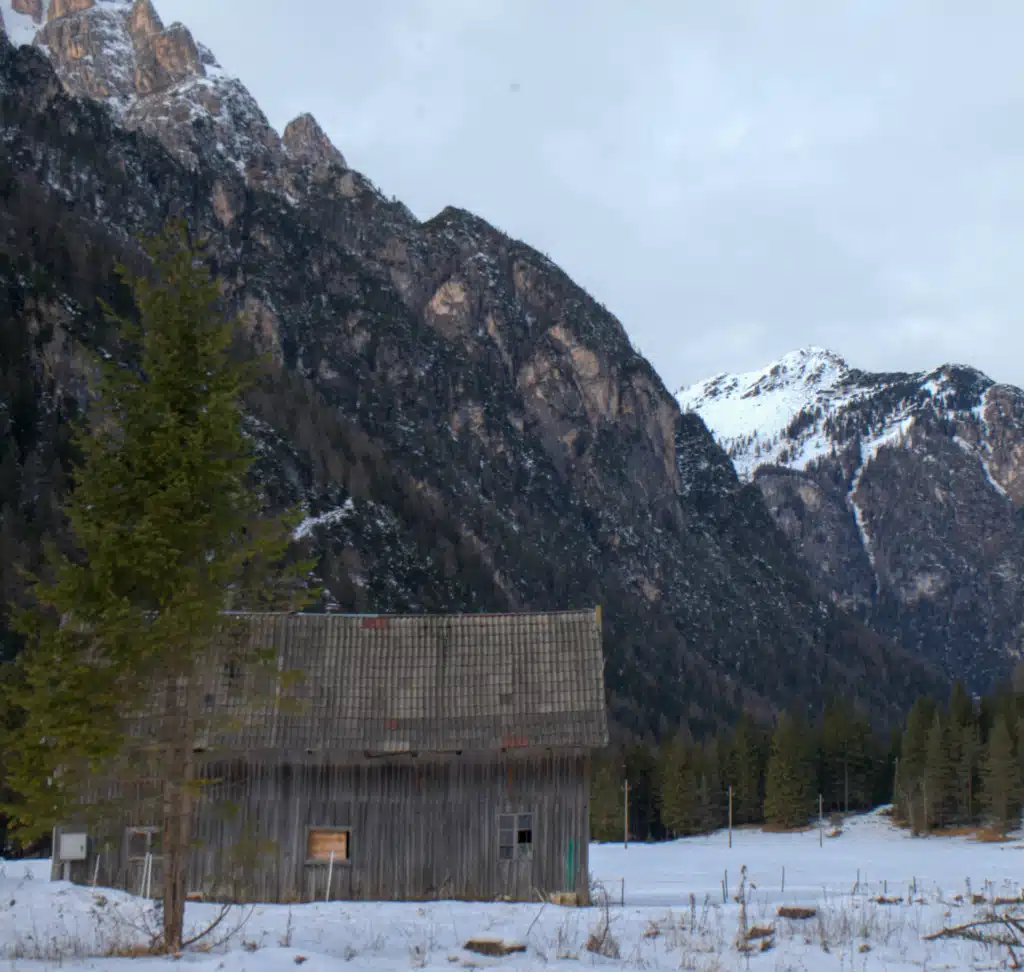 Weathered timber alpine barn in a snowy meadow beneath steep Dolomite mountains in winter, Val Pusteria, South Tyrol.