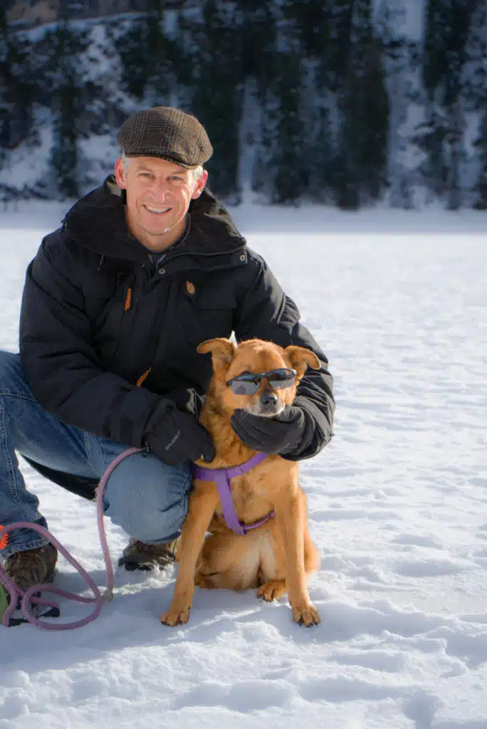 Smiling man kneeling beside a brown dog wearing sunglasses on the snow at Lago di Braies in winter, Dolomites, Italy.