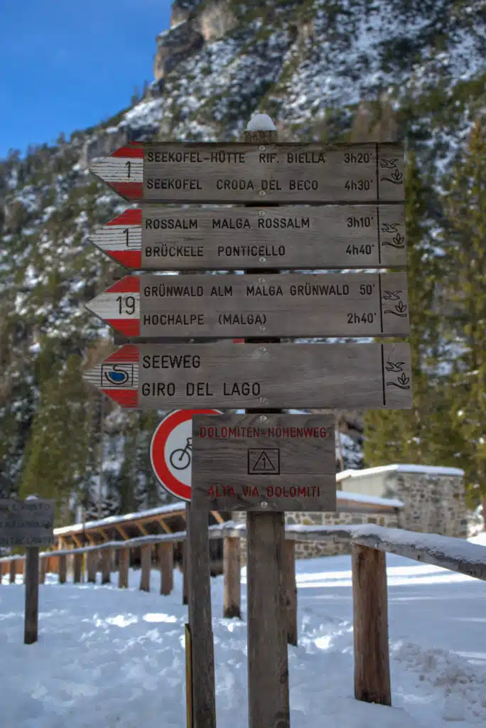 Wooden trail signpost at Lago di Braies showing Croda del Beco, Giro del Lago and Alta Via Dolomiti 1 routes in winter snow.