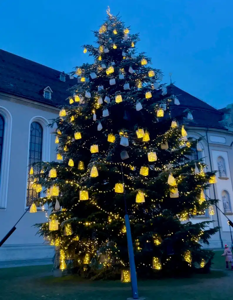 Christmas tree decorated with glowing paper lanterns outside St. Gallen Abbey at dusk