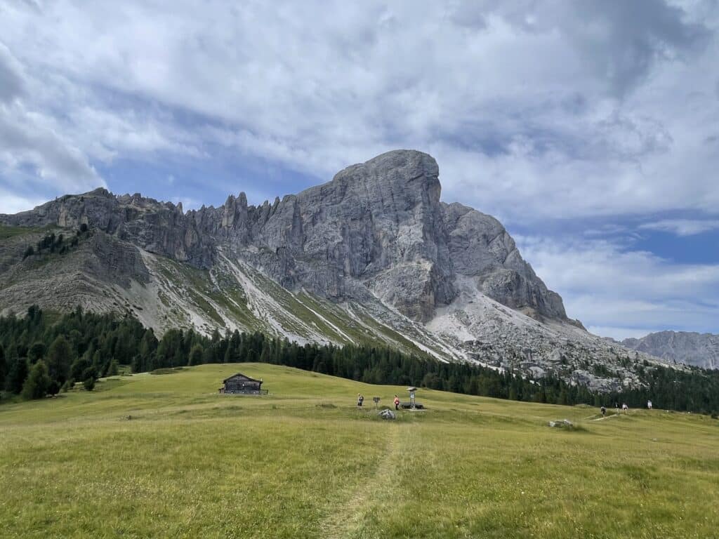 Hikers crossing a wide green meadow with a wooden hut and a large Dolomites cliff face rising in the background