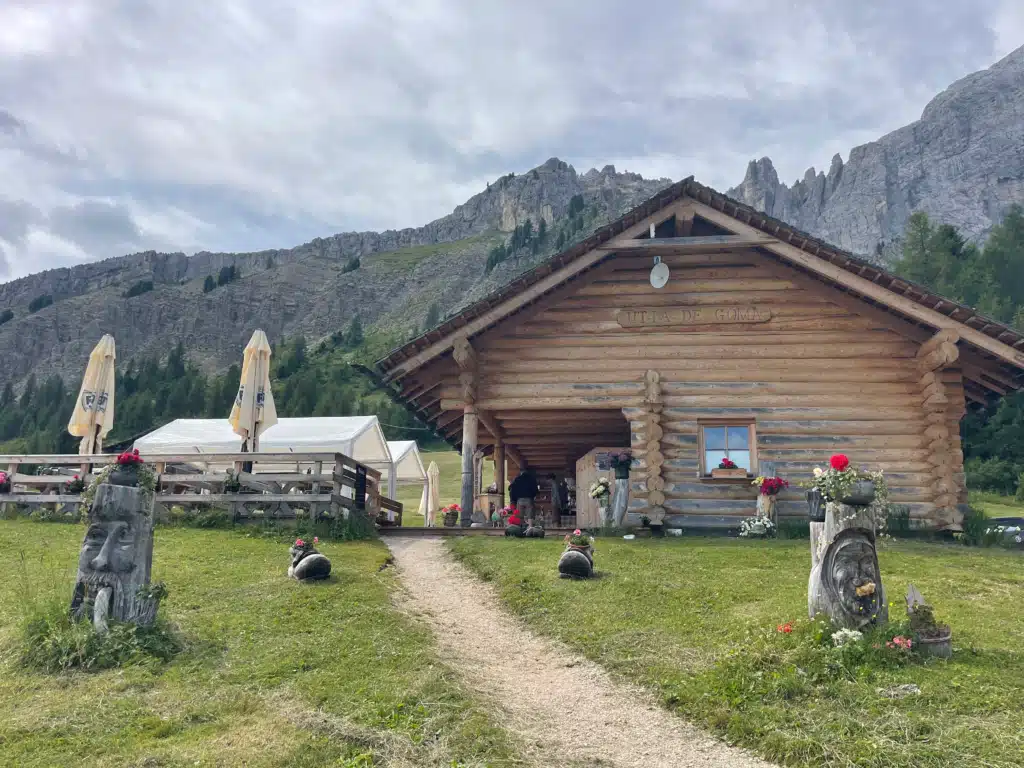Utia de Göma wooden log hut with outdoor seating, carved wooden figures, and jagged Dolomites peaks in the background
