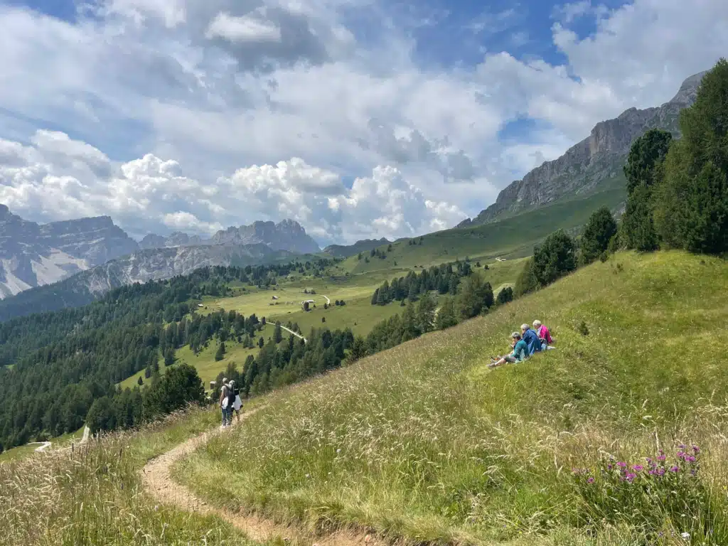 Hikers pausing at Passo Gomo overlooking a green valley with Dolomites peaks and the trail toward Ütia Vaciara