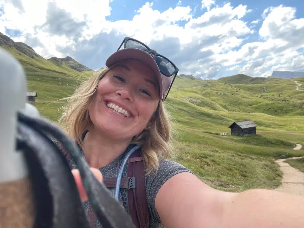 Solo female hiker smiling in a selfie with a wooden hut and rolling green meadow behind her in the Munt de Poz area