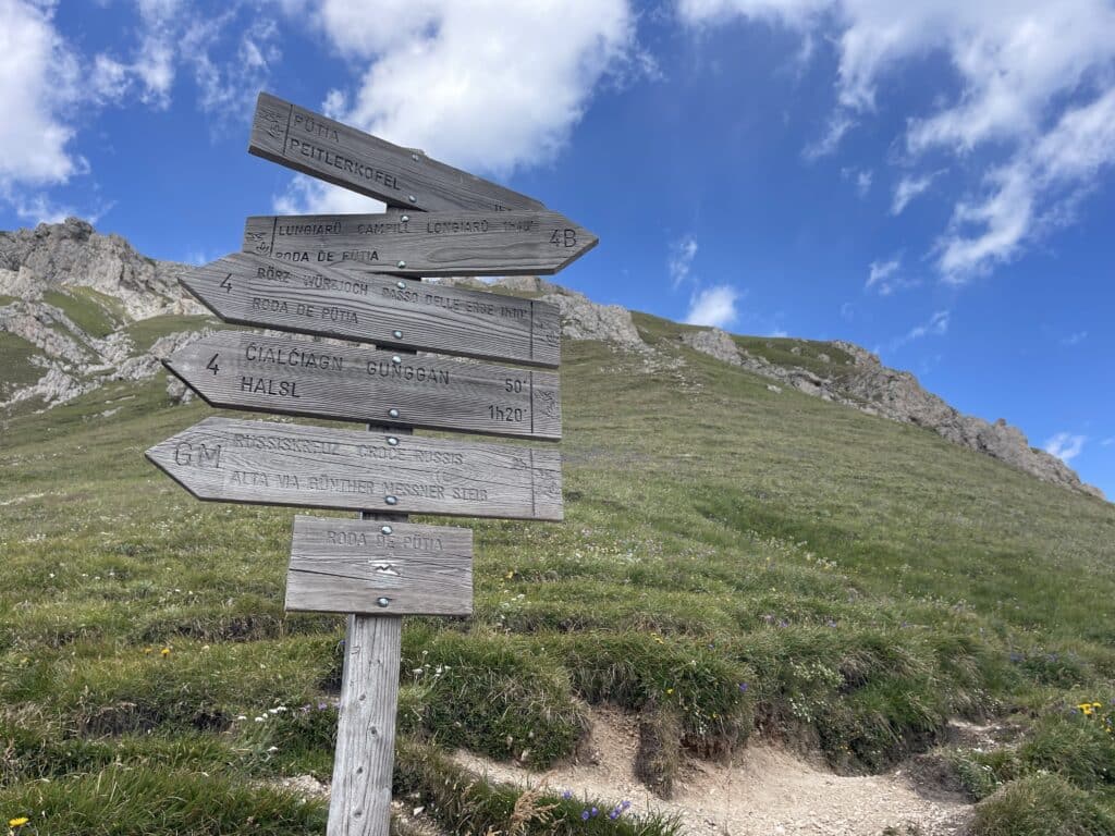 Wooden trail signpost showing multiple routes including Peitlerkofel, Passo delle Erbe, and the Alta Via Günther Messner Steig with hiking times