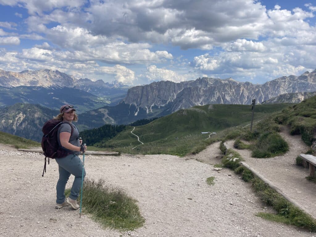 Solo female hiker smiling in a selfie with a wooden hut and rolling green meadow behind her in the Munt de Poz area