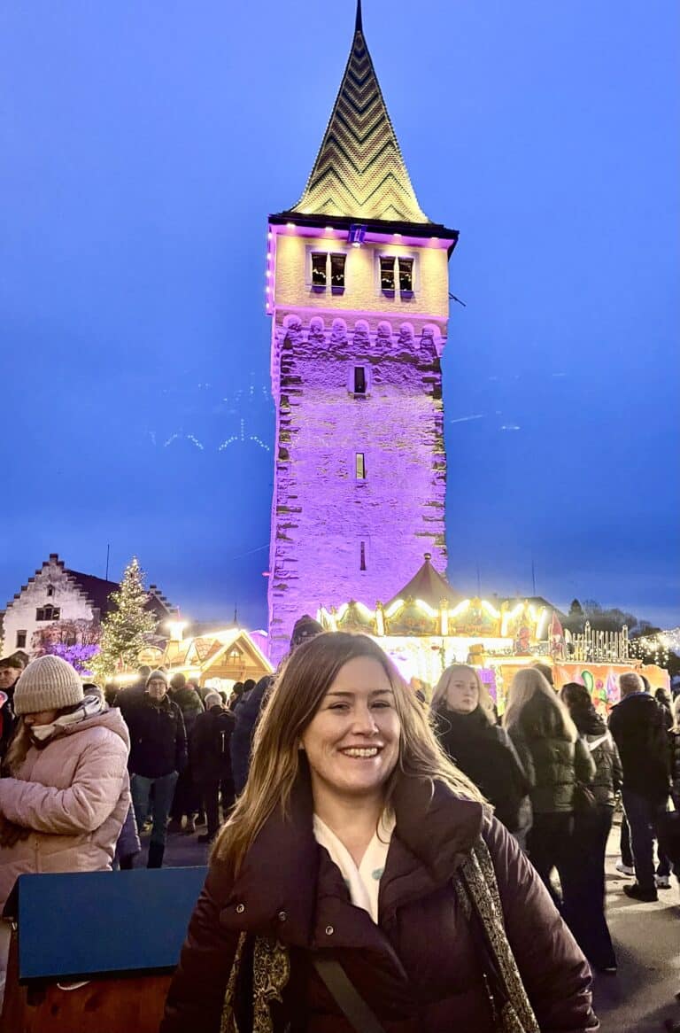 Kimberly smiling at the Lindau Christmas market at blue hour, with the purple-lit Mangturm tower and carousel behind her