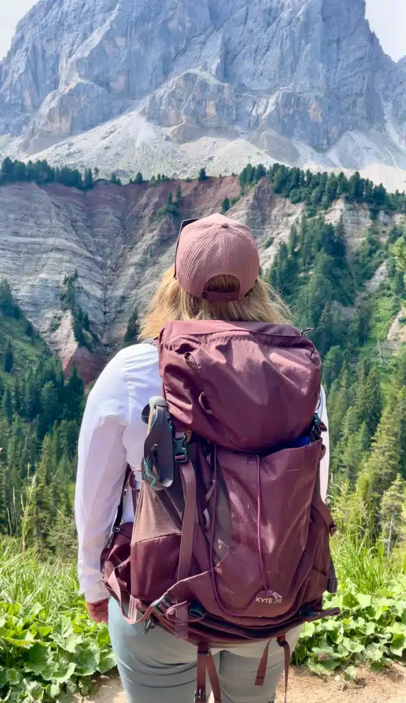 Solo female hiker in a pink cap and white long-sleeve shirt wearing a maroon Osprey Kyte 38 backpack, facing a dramatic Dolomites peak