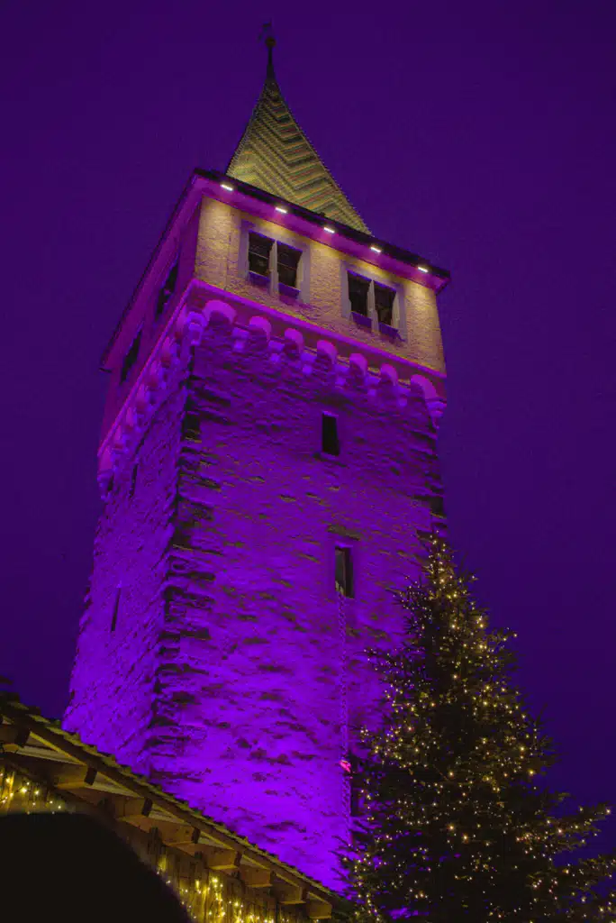 The Mangturm tower lit purple beside a lit Christmas tree at the Lindau Christmas market