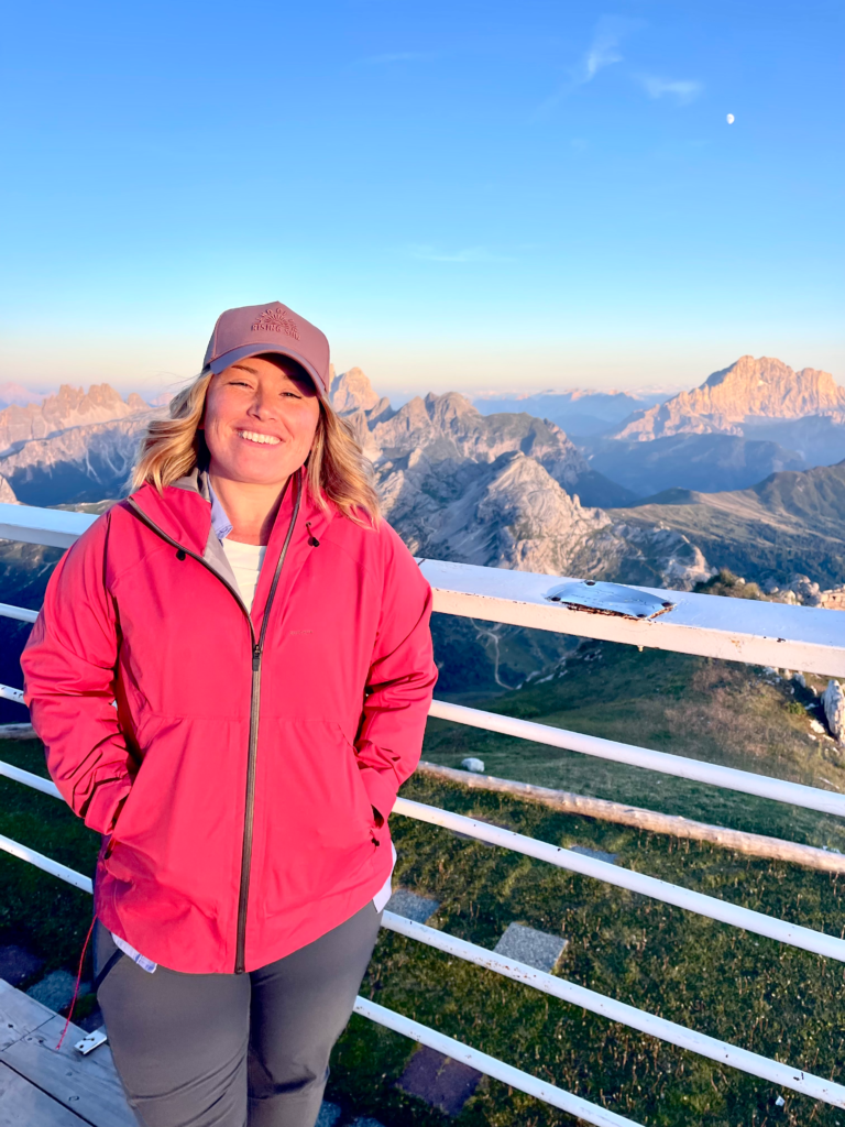 Woman in a red jacket smiling at the railing at Rifugio Lagazuoi with mountains behind her
