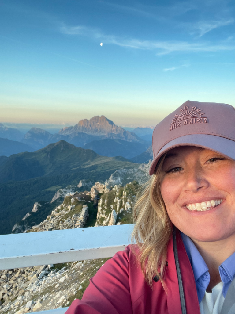 Woman smiling in a pink cap with the Dolomites glowing at sunset behind her