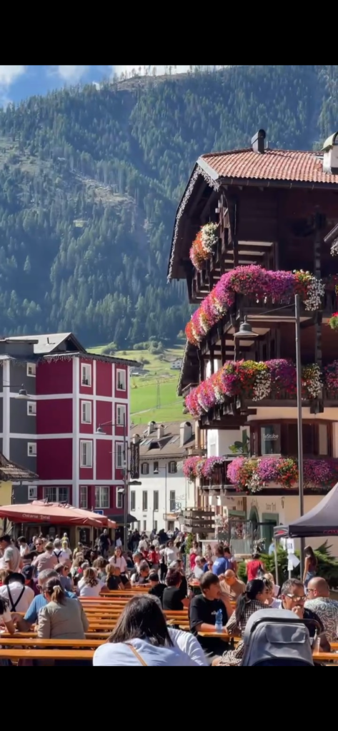 Long communal tables filled with people eating at the Puzzone festival in Moena, flower-decked balconies behind