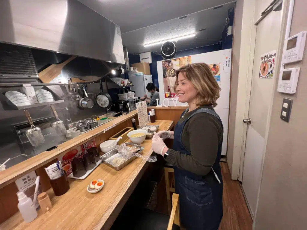 Woman in an apron at the prep counter during a ramen cooking class in Osaka, with noodles and toppings laid out