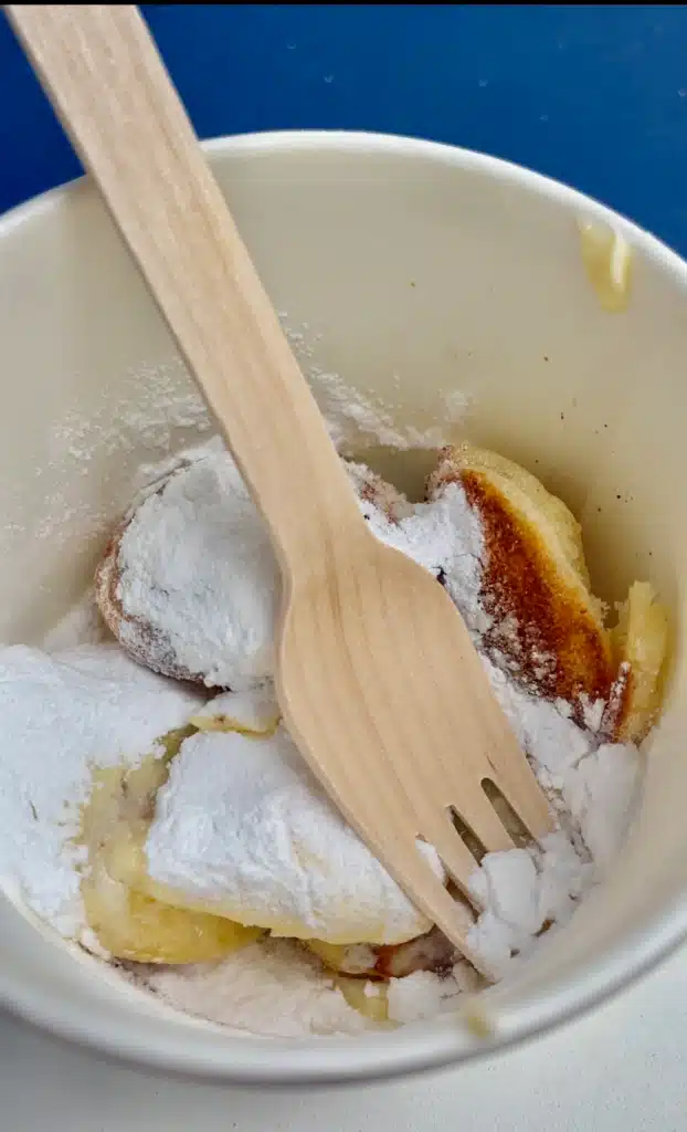 Poffertjes in a paper cup buried under powdered sugar with a wooden fork, eaten on an Amsterdam food tour