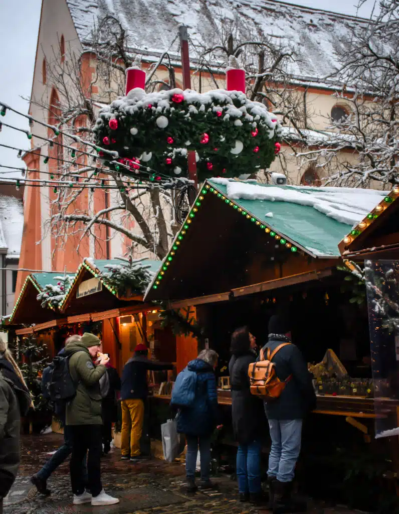 Snow-covered Christmas market stalls under a giant Advent wreath beside a church in Freiburg, Germany