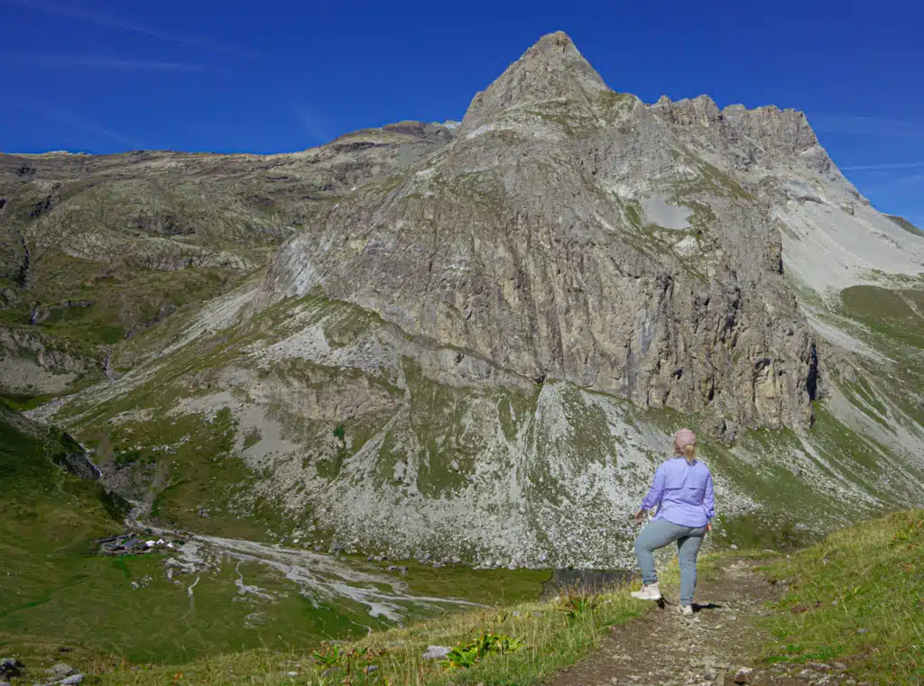 A woman in a purple jacket and pink cap hiking alone on a dirt trail above Refuge Entre-le-Lac, with a sharp rock peak and the Lac de la Plagne valley spread out below her.