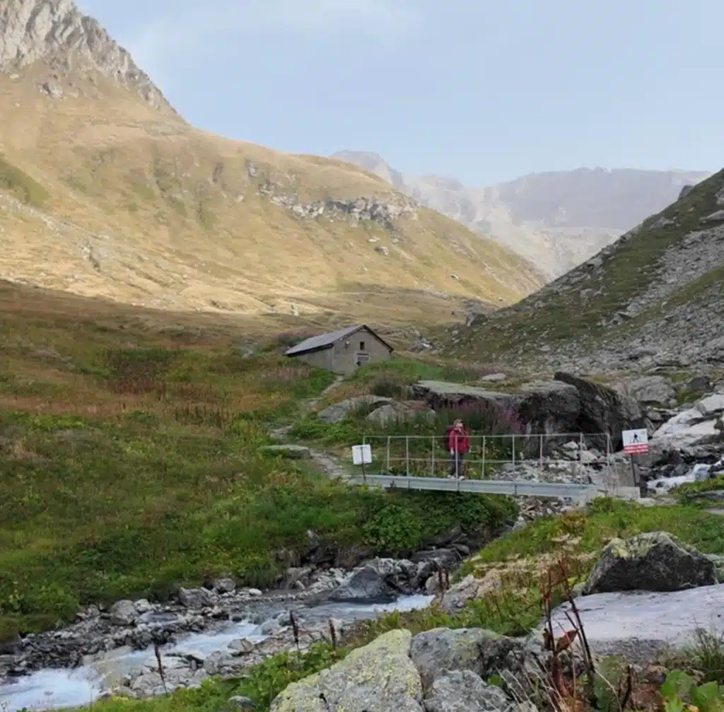Solo female hiker in a red jacket crossing a footbridge over a stream on the Tour du Vallaisonnay in Vanoise National Park