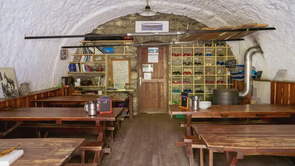 The vaulted stone dining room inside Refuge d'Entre-le-Lac, with long wooden communal tables, a rack of hut shoes sorted by size, shelves of books, and chalkboard signs marking table assignments.