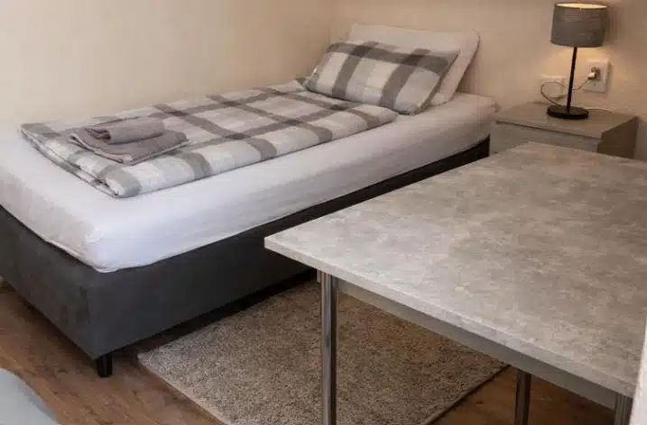 a room with a single bed with grey bedding and a grey table with a lamp at pension bergseeblick in titisee