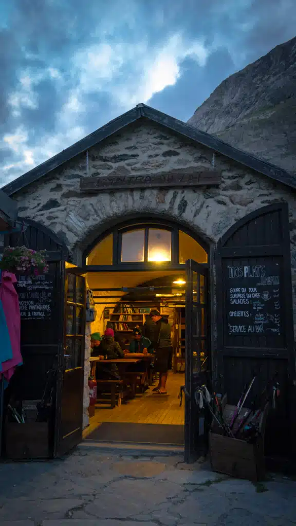The lit stone entrance of Refuge d'Entre-le-Lac at dusk, with hikers sitting at wooden tables inside, chalkboard menus on both doors, and crates of trekking poles stacked outside.