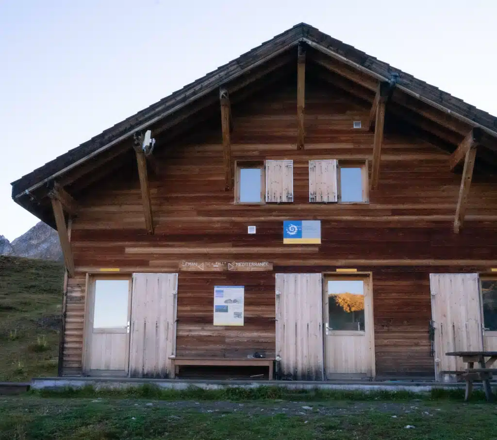 The dark timber front of Refuge du Col du Palet with shuttered windows, a Vanoise National Park sign, and a hand-painted GR5 marker pointing to Lake Geneva in one direction and the Mediterranean in the other.