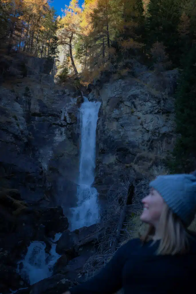 The Cascate Saent waterfall dropping in two tiers through dark rock, framed by golden larch trees, with a timber trail railing in the foreground