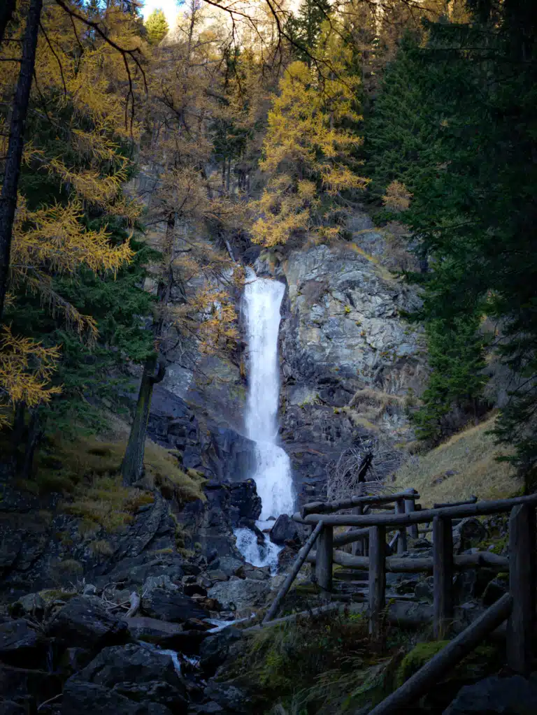 The Cascate Saent waterfall dropping in two tiers through dark rock, framed by golden larch trees, with a timber trail railing in the foreground