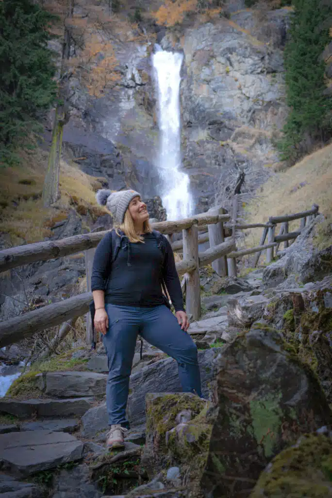 Woman in a knit beanie standing on stone steps below the Cascate Saent waterfall in Val di Rabbi, Italy, with a timber trail railing and larch trees behind her