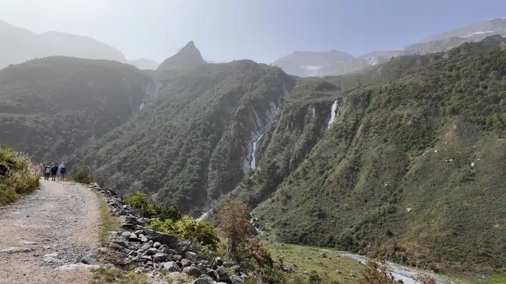 Hikers on a wide gravel path toward Refuge de la Glière with waterfalls on the valley wall, Tour du Vallaisonnay