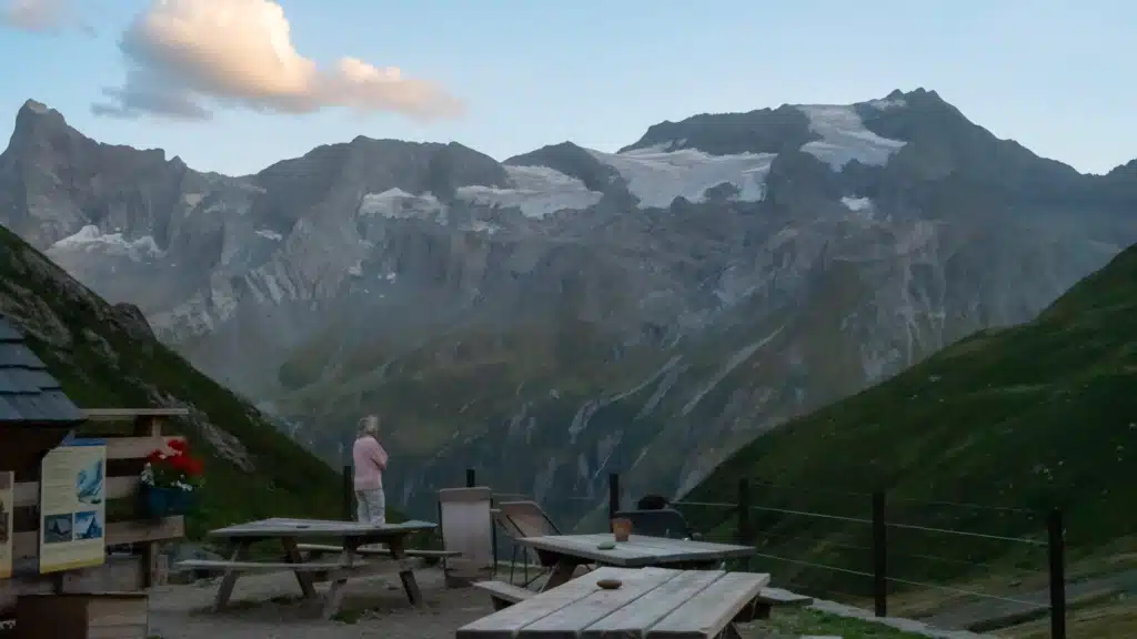 A woman standing on a picnic bench on a refuge terrace at dusk, looking out at a line of glaciated peaks across the valley in Vanoise National Park.