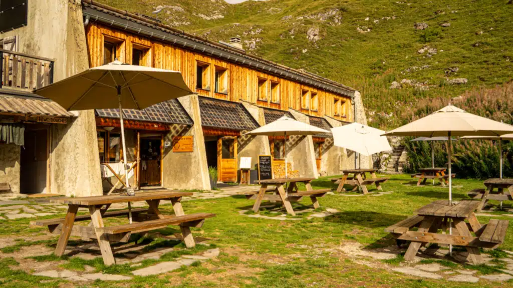 The wooden exterior of Refuge de la Glière in evening light, with picnic tables and cream umbrellas set out on the grass, a chalkboard menu by the door, and a WC and showers sign on the building to the left.