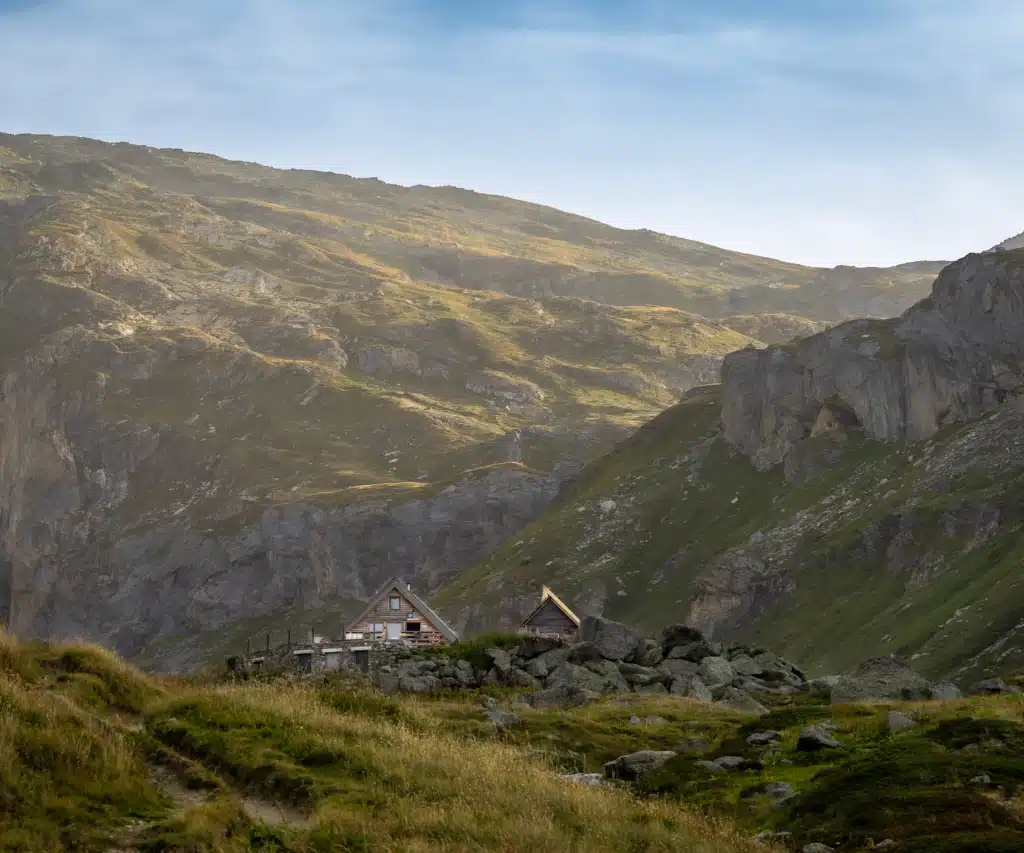 The two wooden chalet buildings of Refuge de Plaisance sitting on a rocky shelf in Vanoise National Park, with a trail running through the grass in the foreground and steep mountain slopes rising behind.