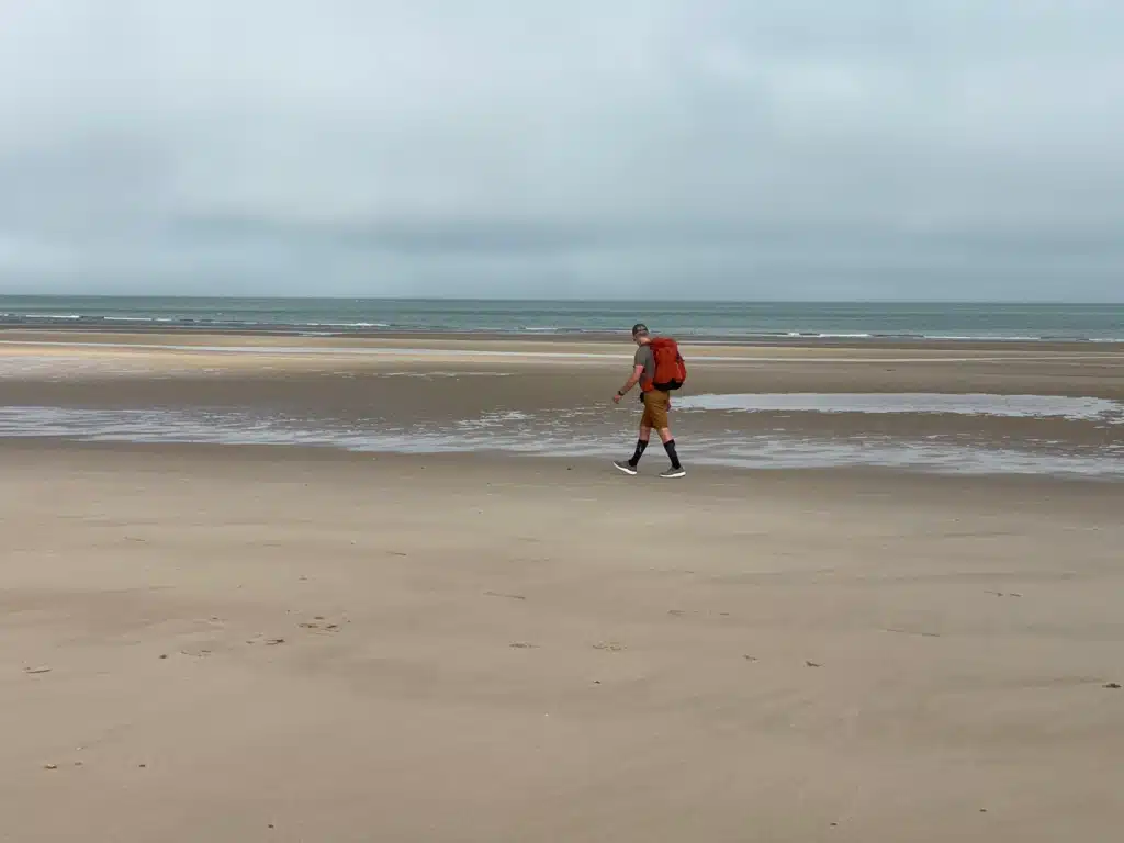Hiker with an orange backpack crossing the wide sandy beach at low tide between Wissant and Tardinghen