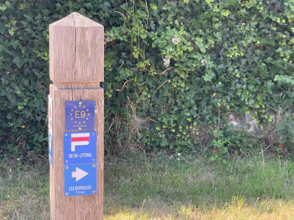 Trail signpost with hiker Trail marker and Cap Gris-Nez information sign on the GR120, with a hiker on the path ahead
